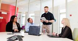 In a conference room, one male presenter is standing while three seated co-workers are listening.